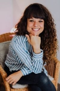 Image of a woman sitting on a rattan armchair, wearing a striped blue and white shirt looking relaxed and smiling at the camera